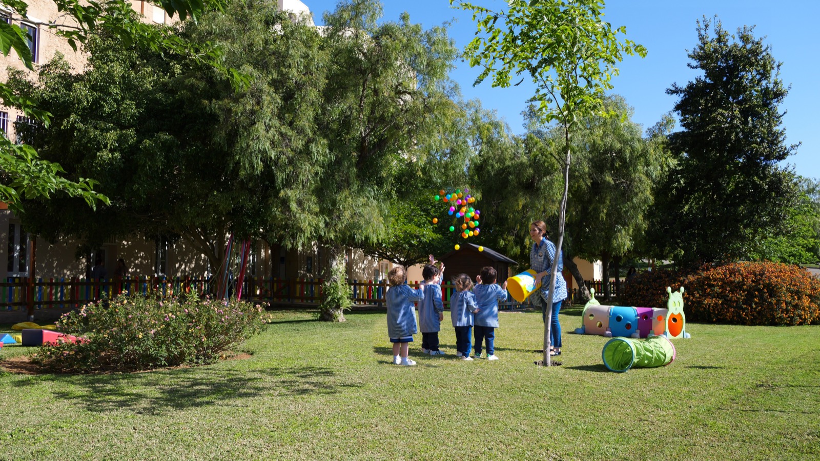 Educadora jugando con pelotas de colores junto a alumnos de la Escoleta en el jardín